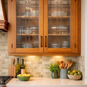 May include: Wooden kitchen cabinets with glass-paned doors. The upper cabinets have a grid design and hold dishes. Below, a countertop displays a cutting board, bottles, a bowl of lemons, and a plant. The lower cabinets have drawers.