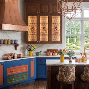 May include: A luxurious kitchen with dark wood cabinetry, blue and orange cabinets, and copper accents. Features include a copper range hood, copper pots, and a geometric chandelier. The kitchen has a marble countertop and a view of a garden.