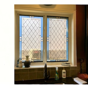 May include: Two stained glass windows with a diamond pattern in a kitchen setting. The windows have a blue border and are set in a white frame. The windows are above a tiled counter with a sink and cleaning supplies.