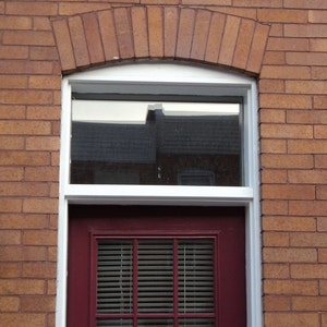May include: A red brick building with a white window and a red door. The window has a white frame and the door has a white frame and a white paneled door with a red door handle. The door has a white window with a white frame and a white paneled door with a red door handle.