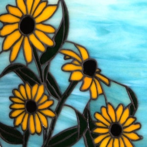 May include: A stained glass artwork featuring four yellow flowers with black centers against a blue background. The flowers are arranged in a cluster, with the largest flower in the upper left corner. The artwork is framed with a black border.