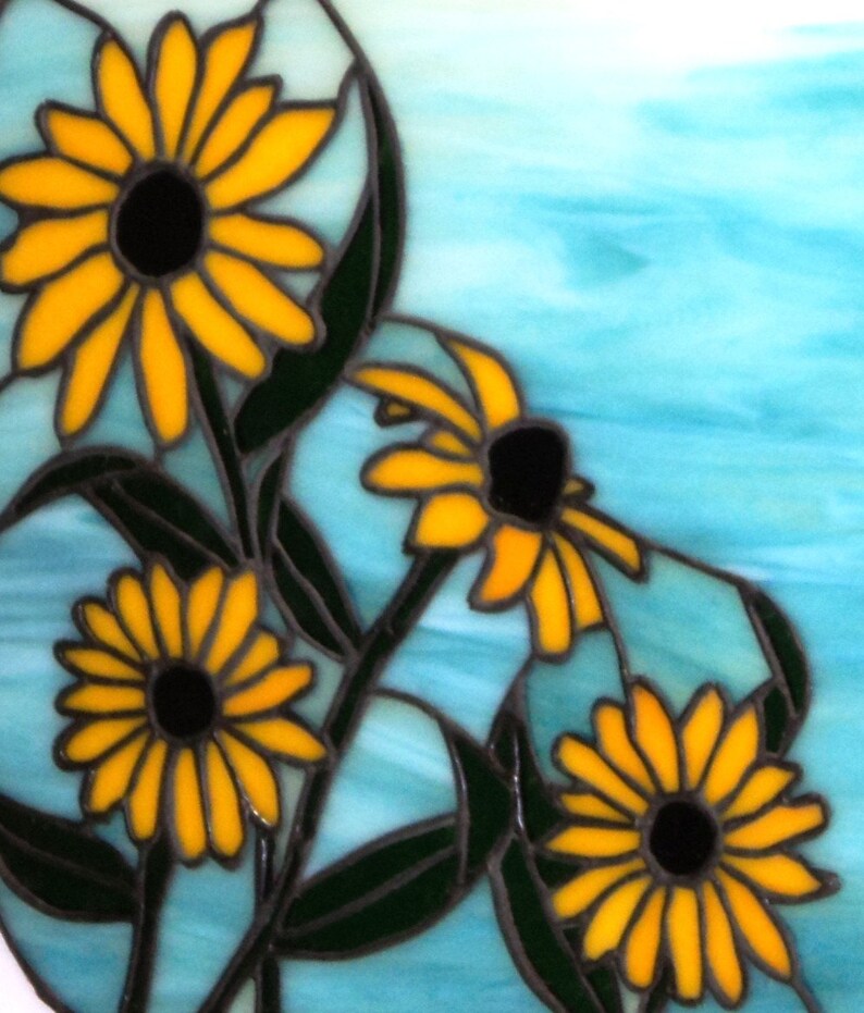 May include: A stained glass artwork featuring four yellow flowers with black centers against a blue background. The flowers are arranged in a cluster, with the largest flower in the upper left corner. The artwork is framed with a black border.