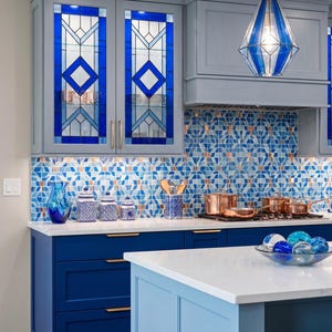 May include: A modern kitchen with blue cabinetry, a white countertop, and a mosaic tile backsplash. The upper cabinets feature stained glass panels with geometric designs. A blue and white pendant light hangs above the island.