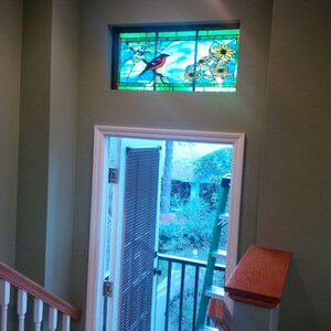 May include: A view from a staircase looking out a door with a stained glass window above. The stained glass window features a bird and flowers. The door is open and leads to a patio with a ladder and a view of trees.