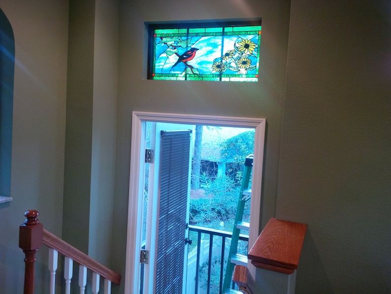 May include: A view from a staircase looking out a door with a stained glass window above. The stained glass window features a bird and flowers. The door is open and leads to a patio with a ladder and a view of trees.