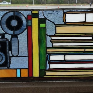 May include: A stained glass window depicting a stack of books, a vintage camera, and a magnifying glass. The books are in various colors, including red, blue, green, and yellow. The camera is black and silver, and the magnifying glass is silver with a black handle.