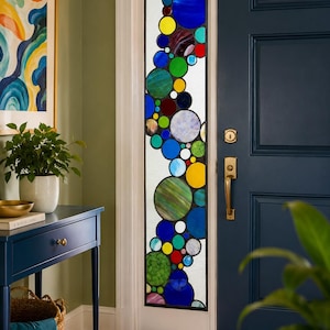 May include: A stained glass window panel with a design of colorful, overlapping circles in shades of blue, green, yellow, and red. The panel is set in a white frame next to a navy blue door. The door has a brass handle.