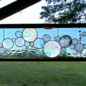 May include: A rectangular stained glass window featuring a design of iridescent, overlapping circles in shades of blue, white, and clear. The circles vary in size and texture, with some appearing smooth and others textured. The window is framed in dark brown wood, with a view of a body of water and green grass visible through the glass.