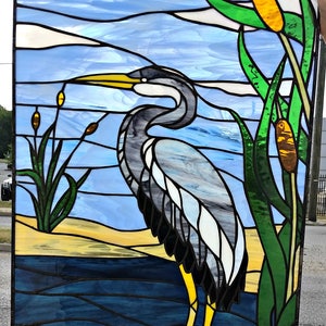 May include: A stained glass window depicting a blue heron standing in a body of water with yellow cattails and green reeds.