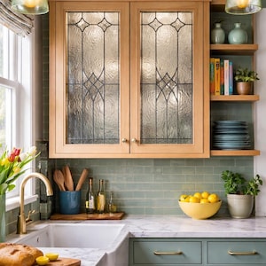 May include: A kitchen with light wood cabinets featuring textured glass doors. The kitchen has a white sink, green cabinets, and a marble countertop. A bowl of lemons sits on the counter, and there are decorative shelves with plants and books.