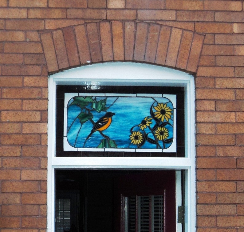 May include: A stained glass window with a blue background, a yellow and orange bird, and yellow flowers. The window is set in a brick wall.