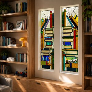 May include: A built-in wooden bookcase with multiple shelves filled with books and decorative items. Two stained glass windows depict stacks of books in vibrant colors. A gray armchair with a colorful pillow is in the foreground.