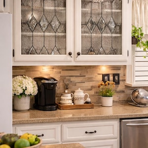 May include: White kitchen cabinets with glass-paneled doors featuring a diamond pattern. A coffee maker, sugar bowl, and teapot sit on the countertop. A bowl of fruit and a striped towel are in the foreground. The kitchen has a neutral color palette.