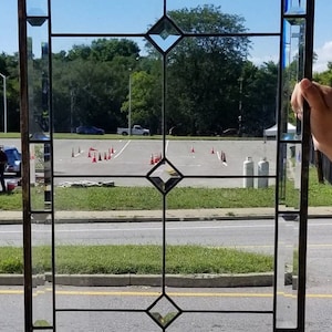 May include: A stained glass window with a diamond pattern. The window is framed in black metal and has clear glass panes. The window is set against a backdrop of a blue sky, green trees, and a paved parking lot.