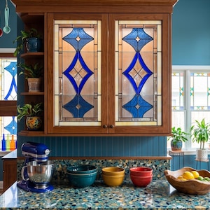 May include: A kitchen with stained glass windows and cabinetry. A blue stand mixer sits on a speckled countertop with colorful bowls. Copper pots and utensils hang above the window.
