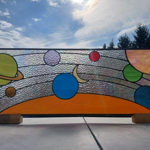 May include: A stained glass panel depicting planets in a solar system design. The planets are various colors, including green, blue, red, and purple, set against a textured, clear background. The panel is framed in wood and features an orange horizon.