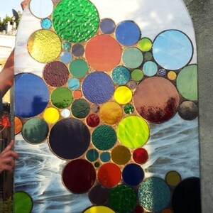 May include: A stained glass window with a design of colorful circles in various sizes. The circles are arranged in a cascading pattern, creating a sense of movement and depth. The window is set against a white background.