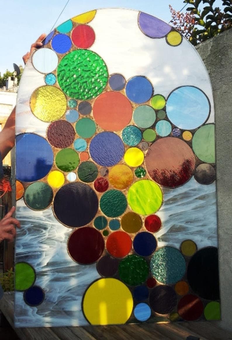 May include: A stained glass window with a design of colorful circles in various sizes. The circles are arranged in a cascading pattern, creating a sense of movement and depth. The window is set against a white background.