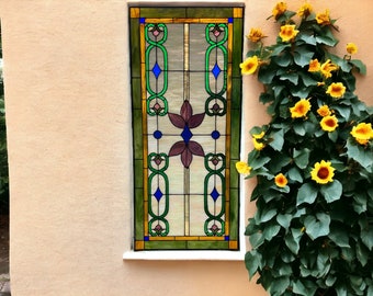 May include: A stained glass window with a geometric design in shades of blue, green, and brown. The window is set in a white frame and is surrounded by a wall with a light beige stucco finish. A vine with yellow flowers grows along the wall to the right of the window.
