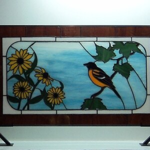 May include: A stained glass window depicting a black and orange bird perched on a branch with green leaves. The bird is surrounded by yellow flowers with black centers. The background is a blue sky with white clouds.