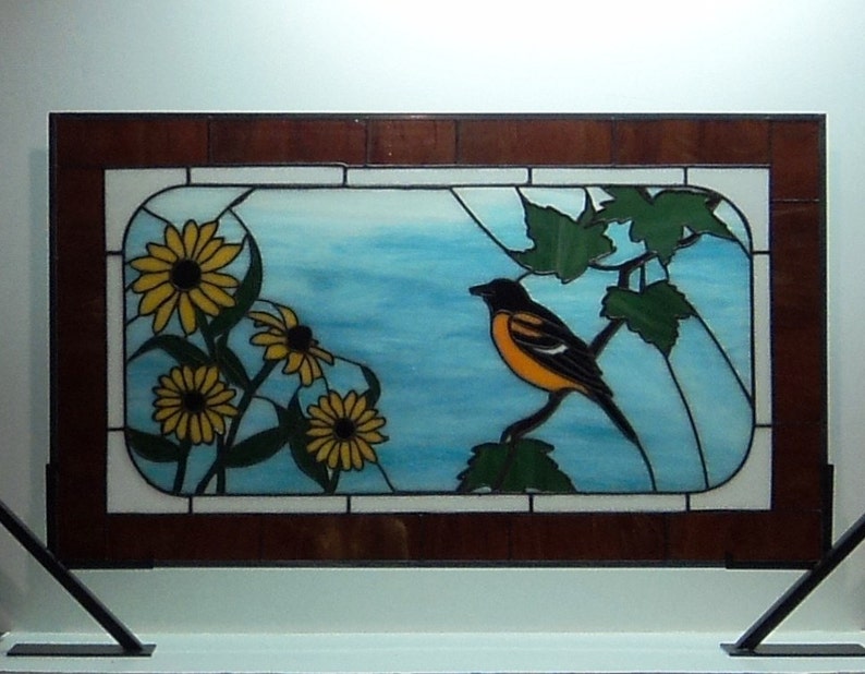 May include: A stained glass window depicting a black and orange bird perched on a branch with green leaves. The bird is surrounded by yellow flowers with black centers. The background is a blue sky with white clouds.