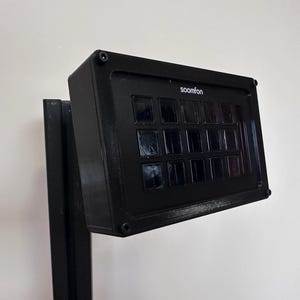 May include: A black electronic device with the word "soomfon" printed on the top. The device has a grid of small, dark squares and is mounted on a black stand. The background is a plain white wall.