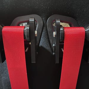 Op de afbeelding: Close-up van een zwarte autostoel met twee felrode veiligheidsgordels. De veiligheidsgordels worden vastgezet door zwarte metalen beugels. De stoel is zwart en heeft een gestructureerd oppervlak. De gordels zijn gemaakt van geweven materiaal.