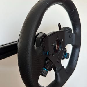 Puede incluir: Un volante de carreras de cuero negro con una superficie texturizada. El volante tiene una variedad de botones y controles, incluido un cambio de marchas azul y plateado.