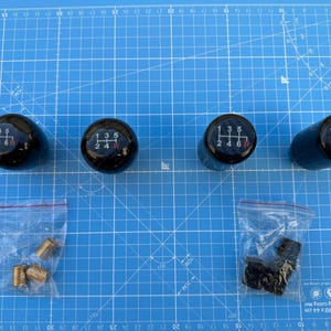 Könnte beinhalten: Vier schwarze Schaltknauf mit weißen Zahlen und einem roten "R" auf einer blauen Schneidematte. Die Knöpfe sind in einer Reihe angeordnet. Auf der Matte befinden sich zwei Beutel mit kleinen Metallteilen.