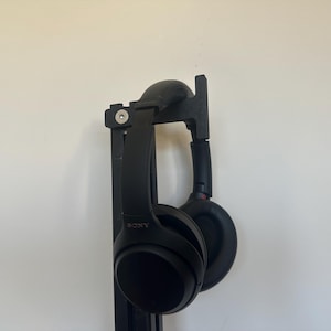 May include: Black over-ear headphones hanging on a black headphone stand. The headphones have a curved headband and large earcups. The stand is mounted on a white wall.
