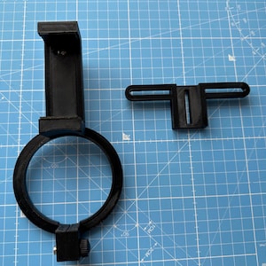 May include: A black plastic phone holder with a ring light attachment. The holder has a clamp to attach to a tripod or other surface. The ring light has a black plastic frame and a white light source.