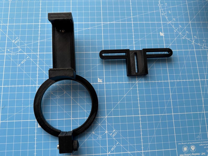 May include: A black plastic phone holder with a ring light attachment. The holder has a clamp to attach to a tripod or other surface. The ring light has a black plastic frame and a white light source.
