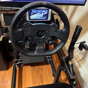 May include: A black racing simulator cockpit with a steering wheel, pedals, and a shifter. The steering wheel has a phone holder with a phone displaying the text "PSR Pure SimRally".