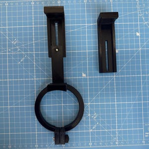 May include: Two black plastic 3D printed parts for a camera mount. The parts are on a blue cutting mat. One part is a ring with a screw on the bottom. The other part is a bracket with a slot for a screw.