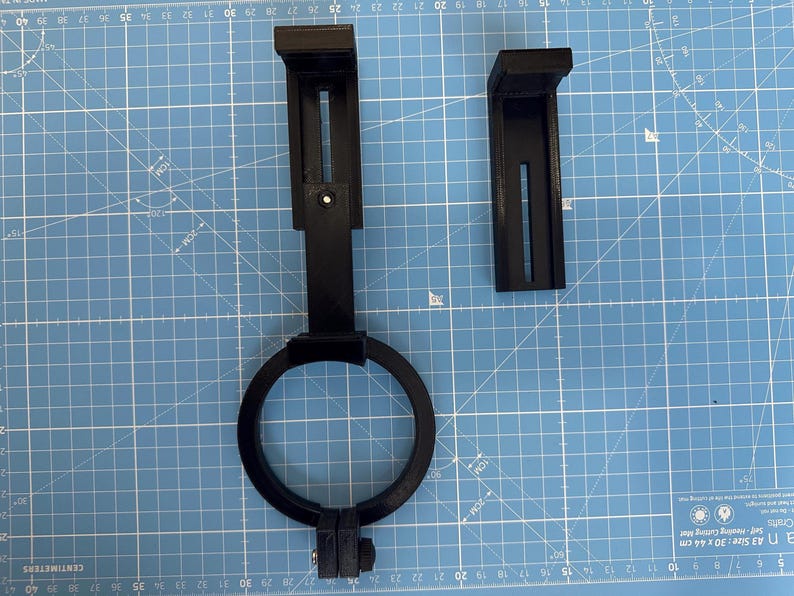 May include: Two black plastic 3D printed parts for a camera mount. The parts are on a blue cutting mat. One part is a ring with a screw on the bottom. The other part is a bracket with a slot for a screw.