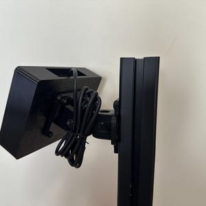May include: A black, rectangular device with a square opening, mounted on a black stand. A bundle of black wires is attached to the device. The stand is made of black metal.
