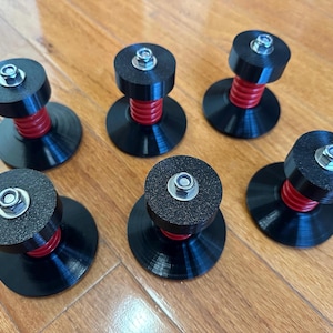 May include: Six black and red speaker isolation feet with a circular base and top, connected by a red spring. Each foot has a silver bolt in the center. The feet are arranged on a wooden surface.
