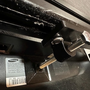 Op de afbeelding: Close-up van een zwarte Samsung TV-beugel met een zilveren schroef en een zwarte moer. De TV-beugel is bevestigd aan de achterkant van een zwarte TV. De TV heeft een label met de tekst "US PAT NO SAT6 STE" en "Contains Mercury, Dispose to Local, State or Federal Regulations". Het label heeft ook een serienummer met de tekst "SIN: CM23H9NS30".