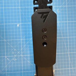 May include: Black metal wall mount with a black plastic plate attached to it. The plate has a logo with the letter "T" in a stylized font. There are three screw holes on the plate.