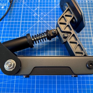 May include: A black and grey racing simulator pedal with a spring and a black cable. The pedal is mounted on a grey base and has a triangular design. The background is a blue cutting mat with a grid pattern.