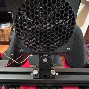 May include: Close-up of a black fan with a honeycomb grill, mounted on a black bracket. The fan is attached to a black and red racing seat with red straps. The image shows the fan's mounting hardware and the seat's frame.