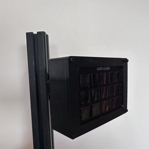 May include: A black electronic device with the word "AMPLIGAME" printed on the front. The device has multiple rectangular buttons and is mounted on a black metal frame. The background is a plain white wall.