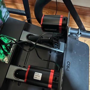 May include: Close-up of a black racing simulator with two red and black pedals and the word "PRO" printed on the side. The pedals are connected to the simulator with black wires.