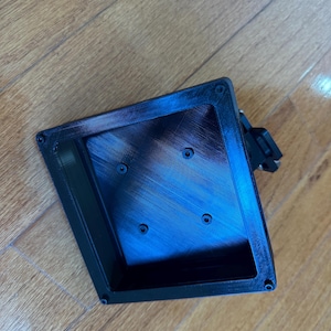 Peut inclure: Un conteneur rectangulaire noir imprimé en 3D avec des coins arrondis et un intérieur texturé. Le conteneur comporte quatre petits trous de vis et un clip noir sur le côté. L'objet est posé sur une surface en bois.