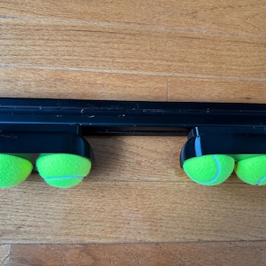 Op de afbeelding: Een zwarte horizontale railbevestiging met twee zwarte kunststof hulpstukken, die elk stevig twee fel neon-groene tennisballen vasthouden. De tennisballen zijn in paren geplaatst, waarbij het geheel een functioneel ontwerp vertoont.