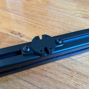 Puede incluir: Un soporte de metal negro con una placa adaptadora redonda de plástico negro. La placa adaptadora tiene una ranura en el centro y cuatro agujeros alrededor del borde. Hay tres tornillos negros que sujetan la placa adaptadora al soporte.