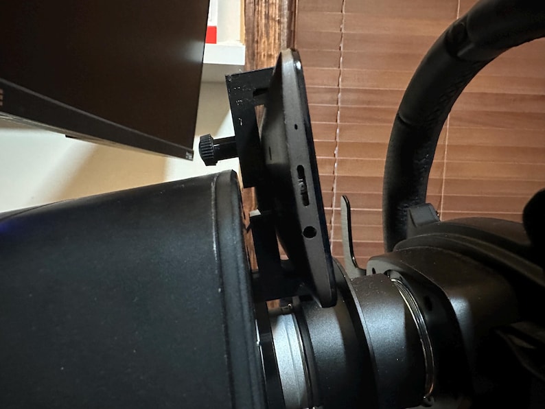 May include: A black steering wheel with a tablet mounted on the side. The tablet is attached to a black mount that is secured to the steering wheel.
