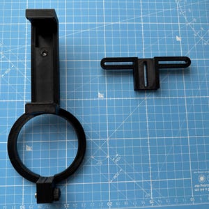 May include: A black plastic phone mount with a ring clamp and a separate black plastic bracket. The mount is designed to attach to a telescope or other optical device.