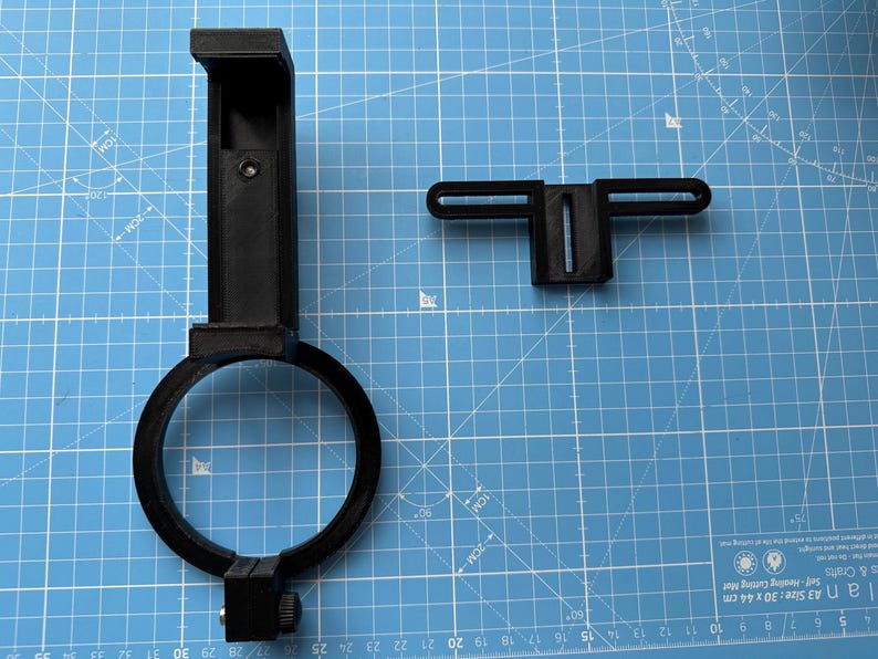 May include: A black plastic phone mount with a ring clamp and a separate black plastic bracket. The mount is designed to attach to a telescope or other optical device.