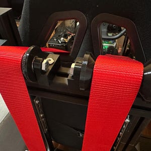 Op de afbeelding: Close-up van een racestoel met een felrode veiligheidsgordel. De gordel is gemaakt van geweven materiaal en wordt vastgezet met zwarte metalen onderdelen. De zwarte stoel maakt deel uit van een racesimulator.